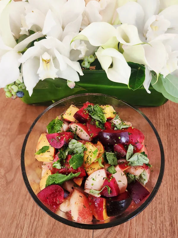 A bowl of colourful fruit beside white flowers on a wooden table.