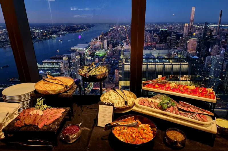 A generous food spread beside windows overlooking a city at dusk.